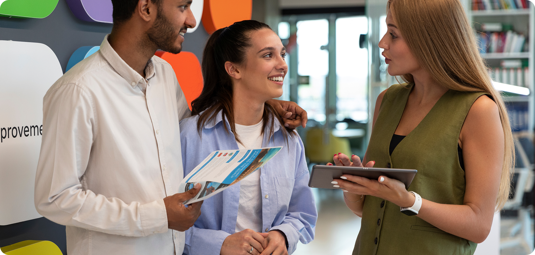 Diverse team chats around office table with laptops and coffee; smiles shared in bright space.